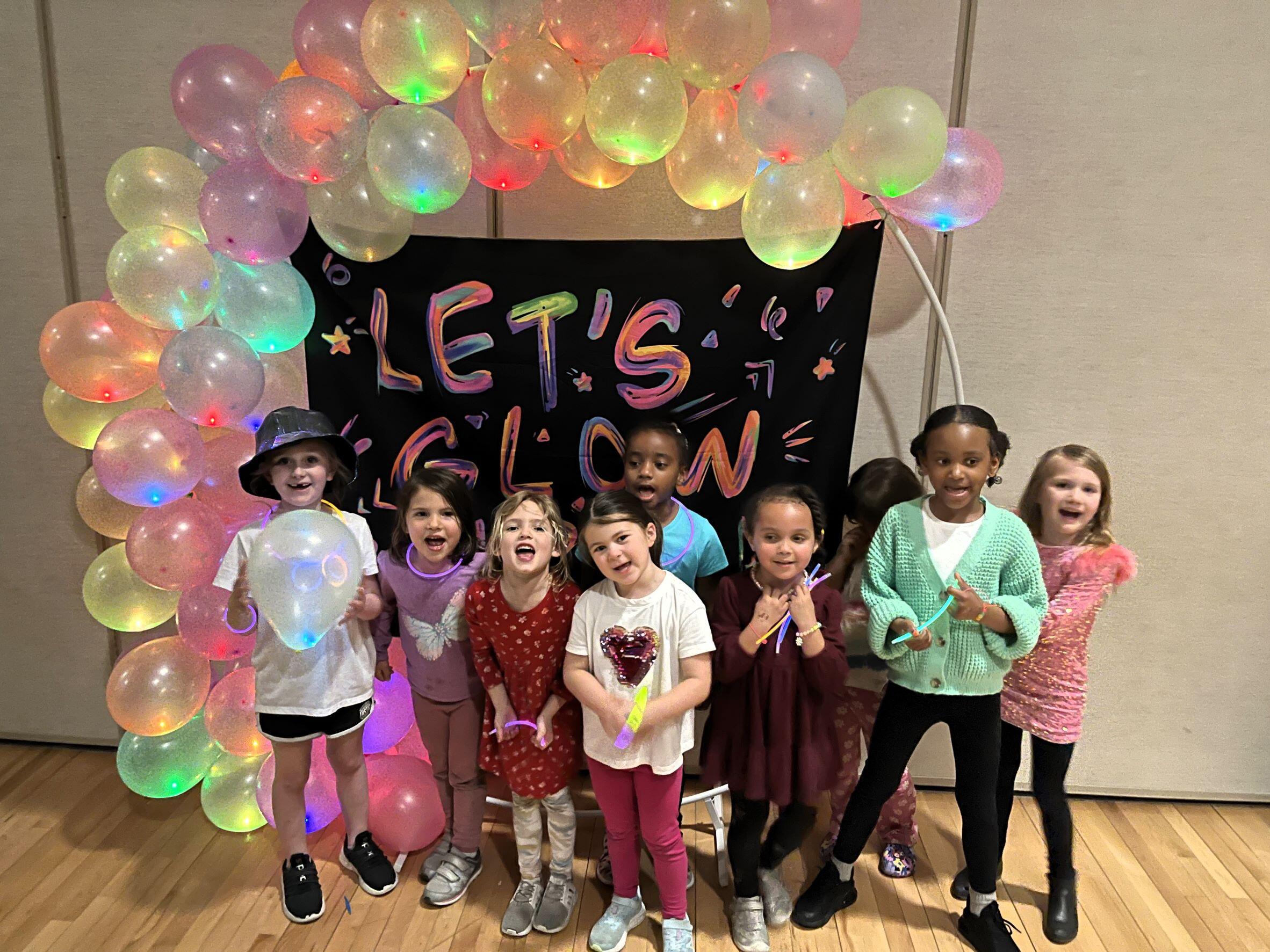A group of 9 kids standing in front of a sign that says Let's Glow. There is a balloon arch behind them. The balloons are glowing all different colors