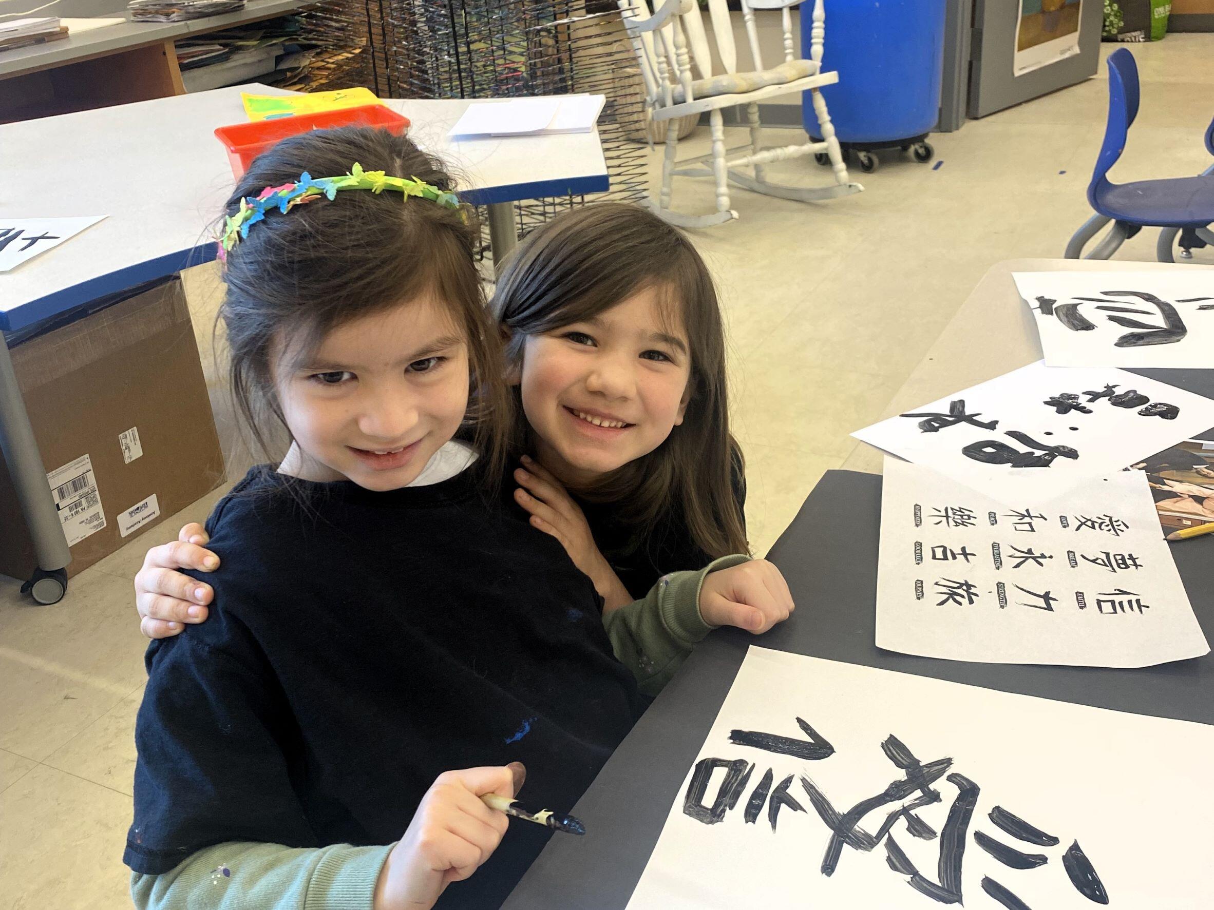 two girls wearing black smocks. They are painting Chinese words on paper.