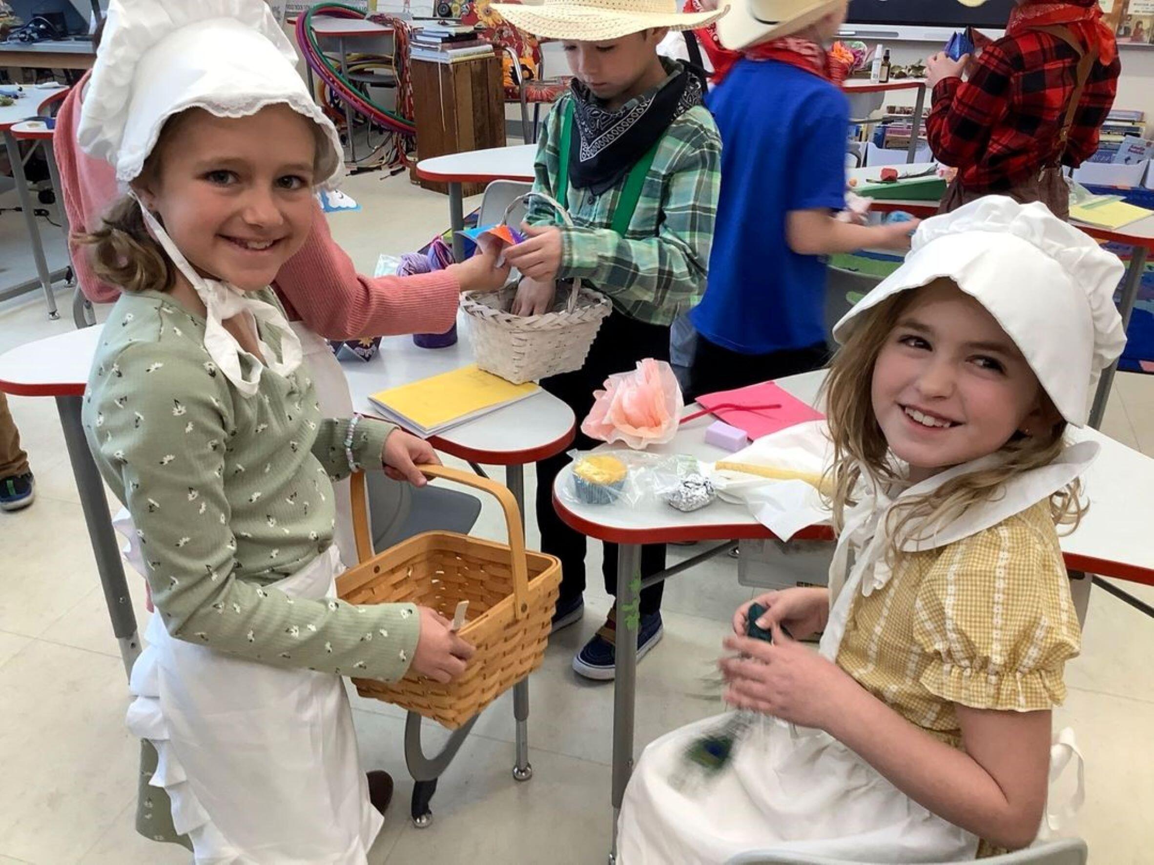 2 female students with bonnets on their heads and aprons on. One is holding a basket the other one has something green in her hands. In the background is a boy wearing a flannel shirt, bandana around his neck and a woven hat. He has his right hand inside of a basket and the left hand is reaching for something that another student who is not visible is holding out.