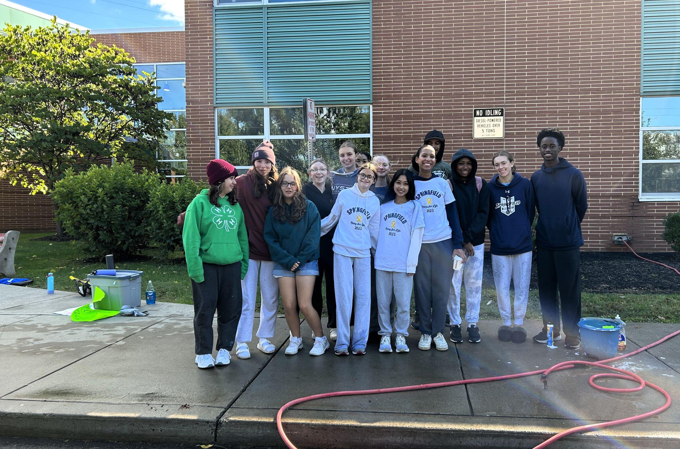 14 Students standing for a group photo. Students had participated in a carwash. There is a hose in front of them on the ground