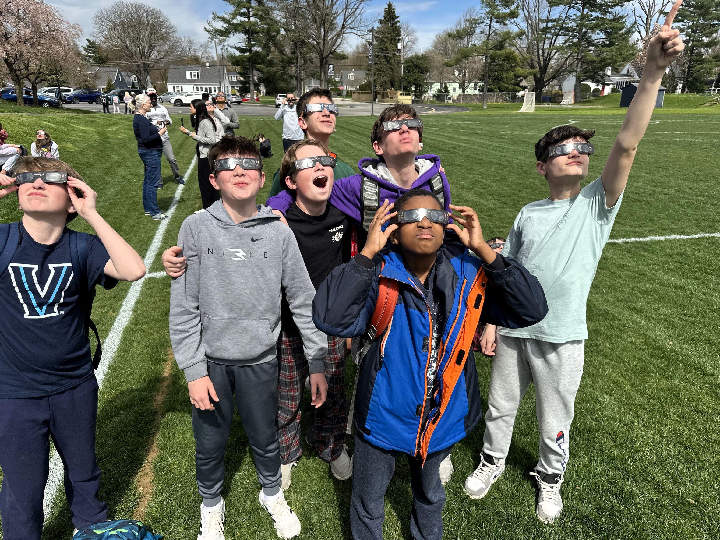 A group of 7 boys standing in a field looking up at the sun, all wearing eclipse glasses. One is pointing up at the sun