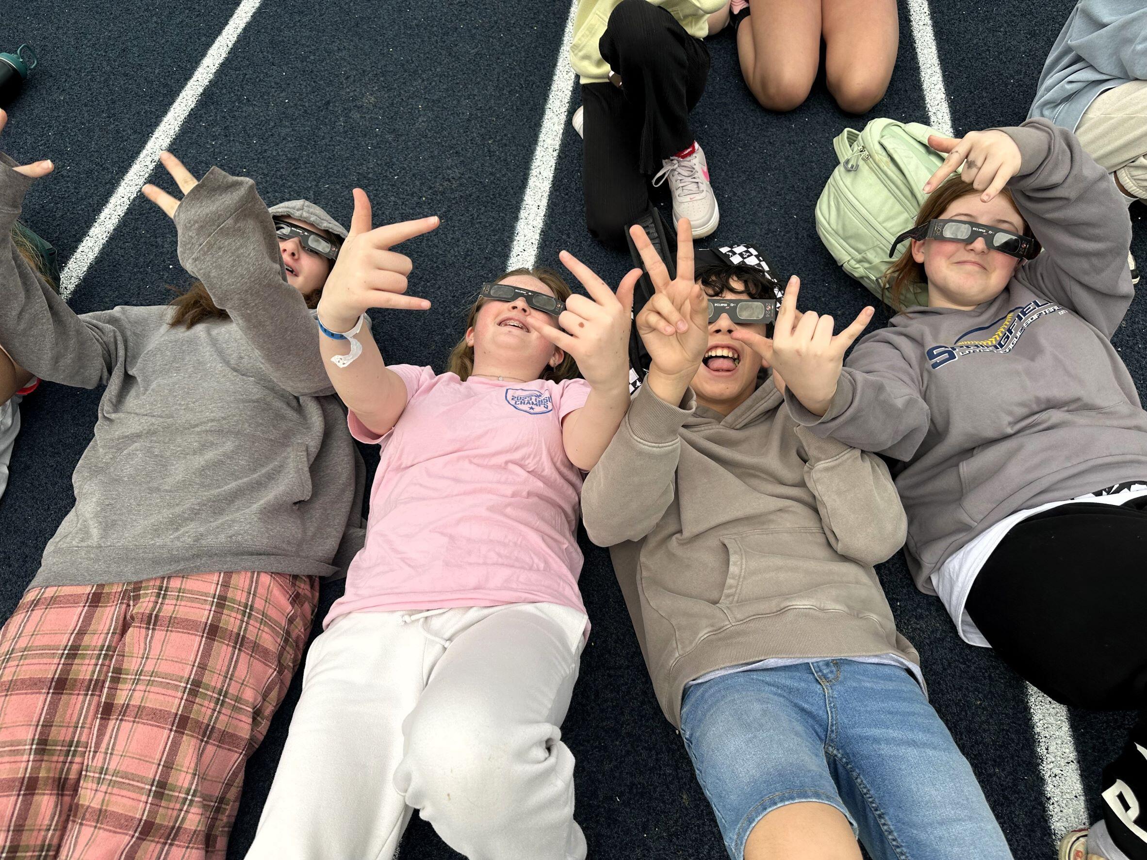 MS- 4 kids laying on the track, wearing eclipse glasses. All kids are smiling at the camera, holding up peace and I love you signs with their hands